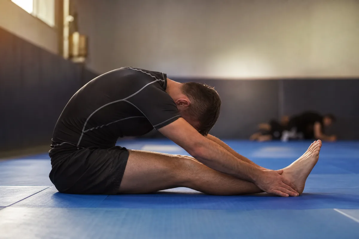 Grappler assis sur un tatami en étirement de la pince, mains tendues vers les orteils, jambes allongées