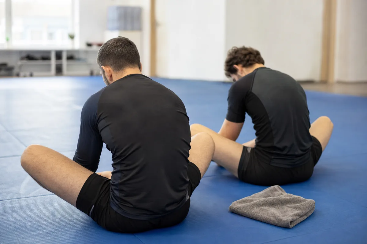 Deux pratiquants en rashguard assis en étirement papillon au bord d’un tatami après l’entraînement